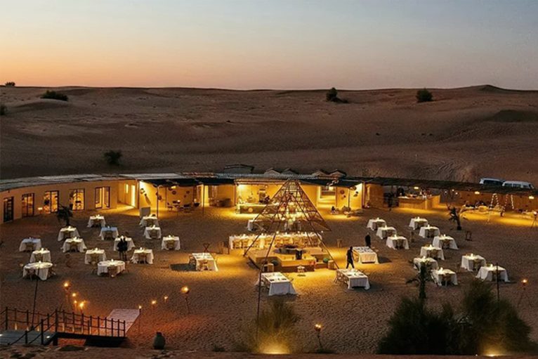 A desert camp of open-sided tents and lanterns on the dunes at dusk